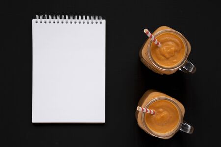 Homemade pumpkin smoothie in glass jars, blank notepad over black background, top view. Flat lay, overhead, from above. Space for text.の写真素材