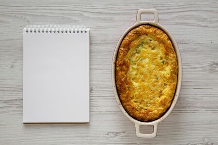 Homemade Cheddar Corn Pudding Casserole, blank notepad on a white wooden background, top view. Flat...