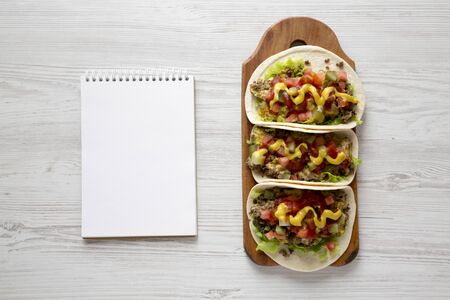 Homemade Cheeseburger Tacos on a rustic wooden board, blank notepad on a white wooden background,...