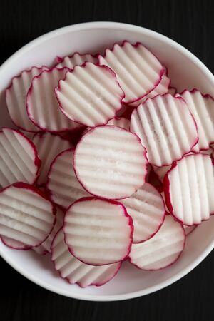 Fresh Radish Chips Slices in a white bowl on a black background, overhead view. Flat lay, top view, from above. Close-up.の写真素材