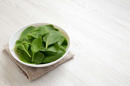 Fresh Baby Spinach in a white bowl on a white wooden background, side view. Copy space.