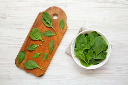 Fresh Baby Spinach in a white bowl on a white wooden table, top view. Overhead, from above, flat...