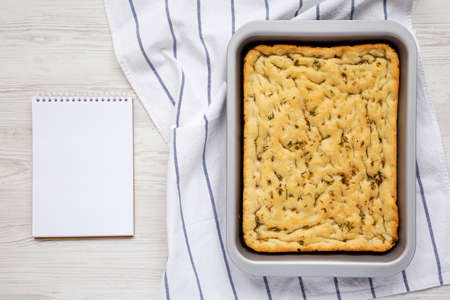 Home-baked Rosemary Garlic Focaccia Bread, blank notepad on a white wooden background, top view....