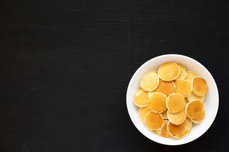 Homemade Mini Pancakes Cereal in a white bowl on a black background, top view. Flat lay, overhead,...