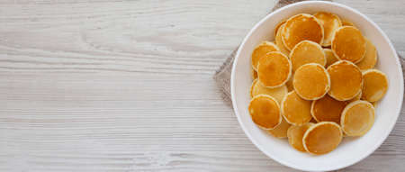 Homemade Mini Pancakes Cereal in a white bowl on a white wooden background, top view. Flat lay,...