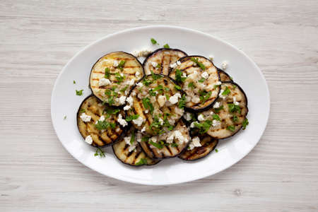 Homemade Grilled Eggplant with Feta and Herbs on a white plate on a white wooden background, top...