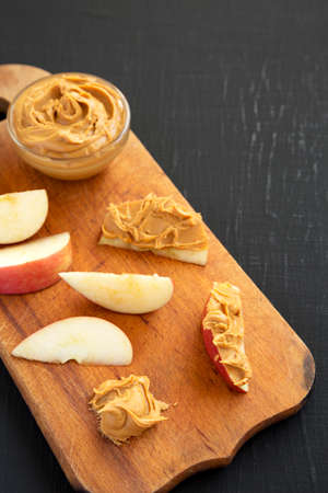 Raw Red Apples and Peanut Butter on a rustic wooden board on a black background, side view. Copy...