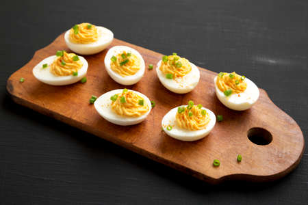 Homemade Deviled Eggs with Chives on a rustic wooden board on a black background, side view.