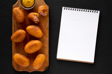 Homemade Mini Corn Dogs on a rustic wooden board, blank notepad on a black background, top view. Flat lay, overhead, from above.の写真素材