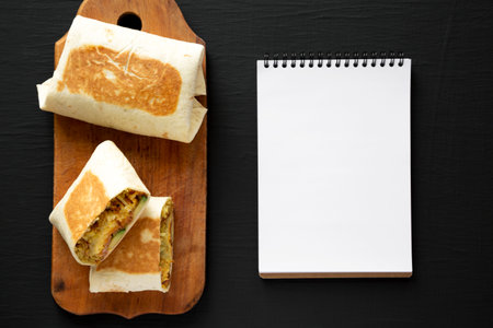 Homemade Breakfast Egg Burrito with Avocado and Bacon on a rustic wooden board, blank notepad on a black background, top view. Flat lay, overhead, from above.の写真素材