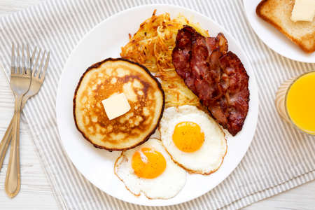 Full American Breakfast with Bacon, Hash Browns, Eggs and Pancakes on a plate on a white wooden...