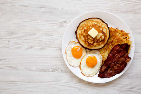 Full American Breakfast with Bacon, Hash Browns, Eggs and Pancakes on a plate on a white wooden...