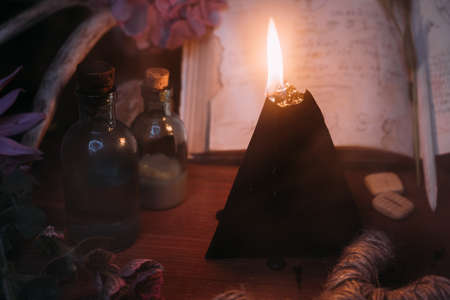 Open old book with magic spells, runes, black candle and herbs on witch table. Occult, esoteric,...