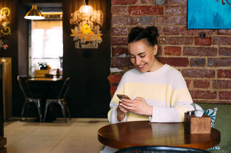 A young girl at a table in a cafe looks at the phone, smiles and texting.
