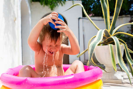 Cute child has fun in inflatable pool with toys in summer in backyard.