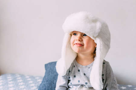 Cute child y on the bed in cozy adult clothes and a hat with earflaps.の写真素材