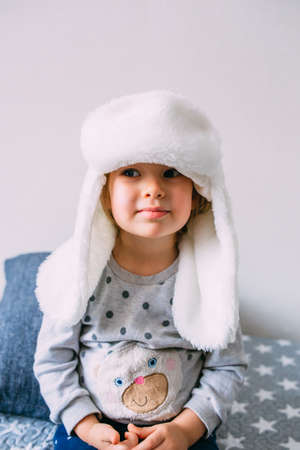 Cute child y on the bed in cozy adult clothes and a hat with earflaps.の写真素材