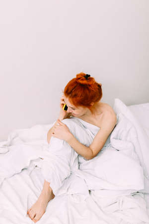 Portrait of a young beautiful redhead woman talking on the phone in bedの写真素材