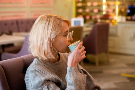 Young attractive blonde girl in cafe drinks coffee from a paper cup and relaxesの写真素材
