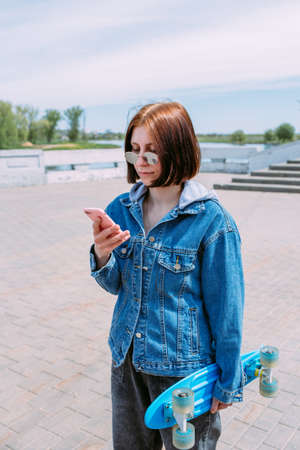 Young stylish woman walks on street with a skateboard and uses a smartphoneの写真素材