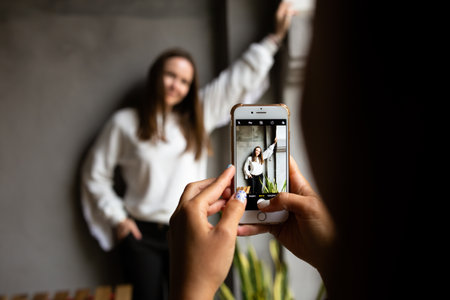 Two young women take photos and videos of each other on smartphone on a terraceの写真素材