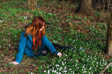 Young red-haired woman sits in the forest among snowdrops with nature