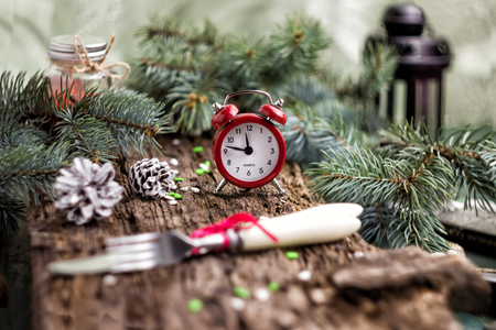 Christmas concept. Alarm clock with spruce pine twigs cones and Cutlery fork and knife on wooden old background. The time of the gala dinner.の写真素材