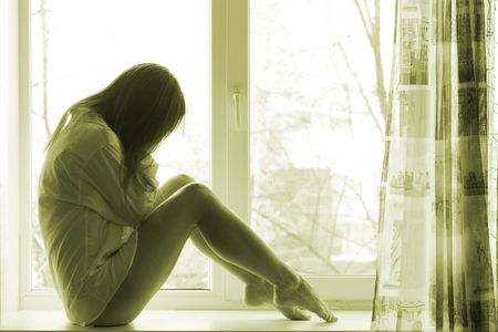 Cute sad girl near window at home in white men shirt, cryingの写真素材