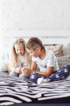 girl and boy sitting in pajamas on the bed and drink milk from a glass cupの写真素材