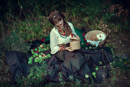 Slavic woman in ethnic dress on grass with apples and basket with bagelsの写真素材
