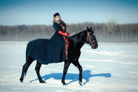 blond woman in red dress with long black coat with dark horse on the background of winter landscapeの写真素材