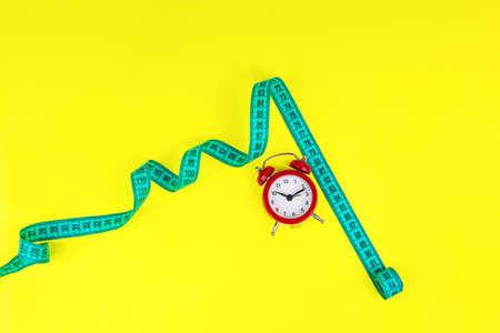 Red alarm clock and bright tape measure on yellow background with copy space