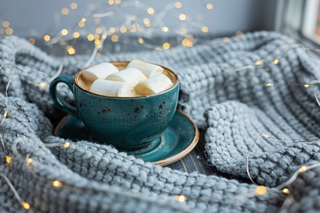 Cup of coffee, marshmallow, warm knitted sweater on wooden background. Warm lights. Cozy winter morning. Lifestyle concept. Selective focus.の写真素材
