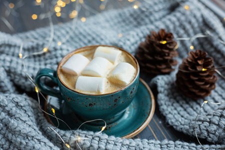 Cup of coffee, marshmallow, warm knitted sweater on wooden background. Warm lights. Cozy winter morning. Lifestyle concept. Selective focus.の写真素材