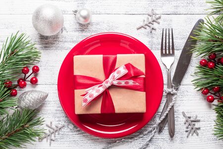 Christmas table setting with red plate, gift box and silverware on light wood background. Fir tree branch, holly berries. Top view with copy space.の写真素材