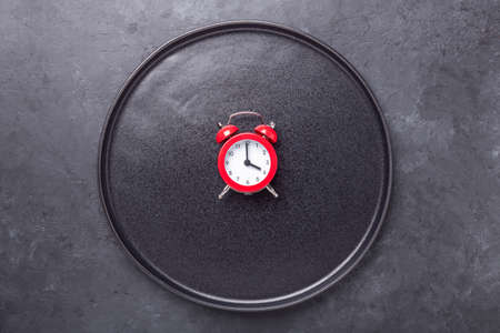 Red alarm clock and empty black ceramic plate on dark stone background. Intermittent fasting concept - Imageの写真素材