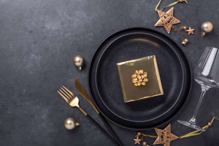 Christmas table setting with black ceramic plate, glass, gift box and gold accessories on black...