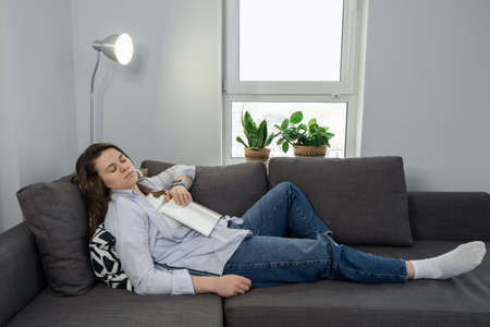 Tired woman napping on sofa at home with book on her chest, taking a break. Fatigue and sleep disorder conceptの写真素材