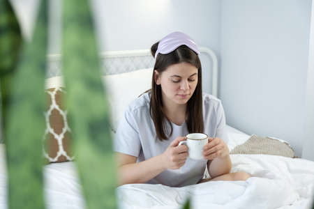 Attractive woman in silk mask having a tea on bed. Portrait of girl enjoying her drink while relaxing at homeの写真素材