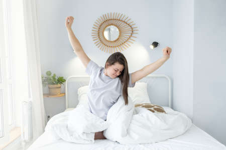 Happy cheerful girl sitting on bed after good sleep. Young woman enjoying morning on bed at homeの写真素材