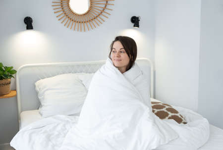 Smiling girl covered with blanket sitting on bed. Young woman enjoying morning on bed at homeの写真素材