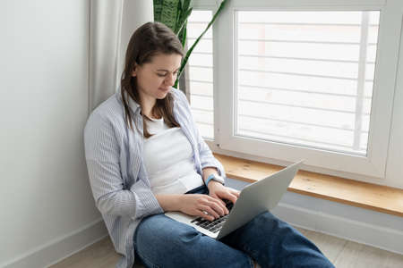 Middle-aged woman working from home. Girl using laptop at home, sitting near a large window. Remote work conceptの写真素材