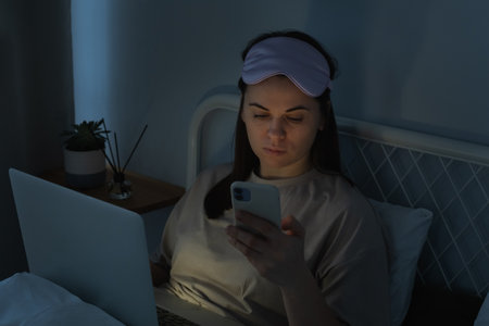 Woman in silk mask using laptop computer and surfing social media on smartphone in bed before sleep at night. Social media addiction conceptの写真素材