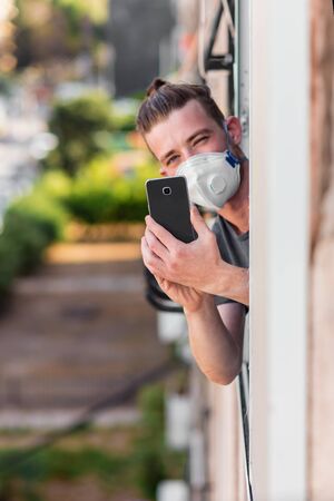 Young man in a medical mask looks out of the apartment window and photographs a neighbor using a mobile phone. Quarantined during the coronavirus epidemicの写真素材