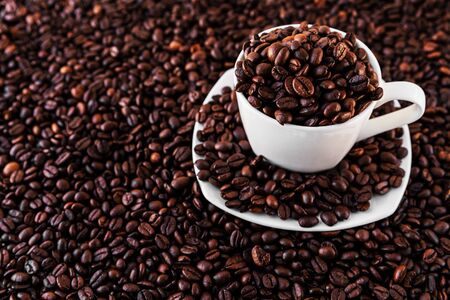 White mug and saucer next to roasted coffee beans everywhere, inside the mug and a lot around it....