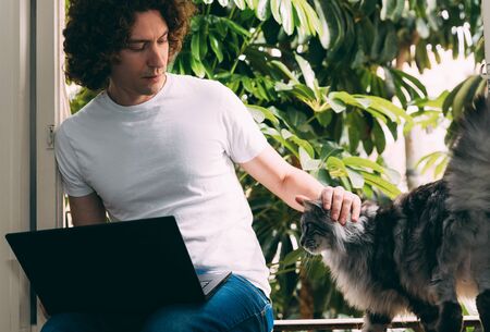 A young curly-haired man in a white T-shirt sits at a window with a laptop and works from home during the quarantine period from coronavirus. He strokes the cat. Outside the window green palm treesの写真素材