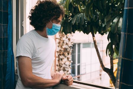 A young curly-haired man in a medical mask and a white T-shirt looks out of the window of the house...