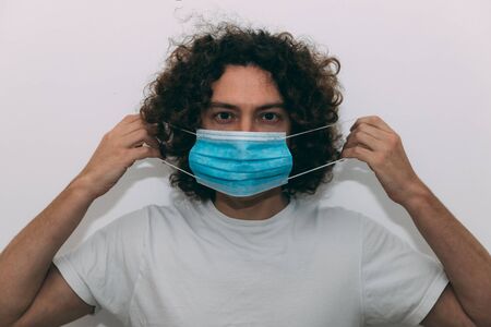 Young man with curly hair in white T-shirt, puts a medical mask on his face. Coronavirus epidemic. Protection of the upper respiratory tract from infection. New reality. Risk of infection. New rules.の写真素材