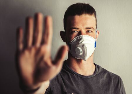 Young man with hand on foreground in a modern medical mask in a gray t-shirt on a light background, during a home quarantine of the coronavirus infection epidemic. Stay home concept. Stop panic.の写真素材