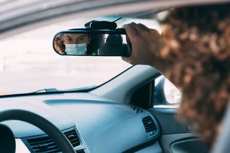 Reflection in the mirror of a man in a medical mask inside a car. Man in protective mask is driving a car. Taxi driver in a mask. Protection from coronavirus epidemic pandemic, covid-19. New realityの写真素材